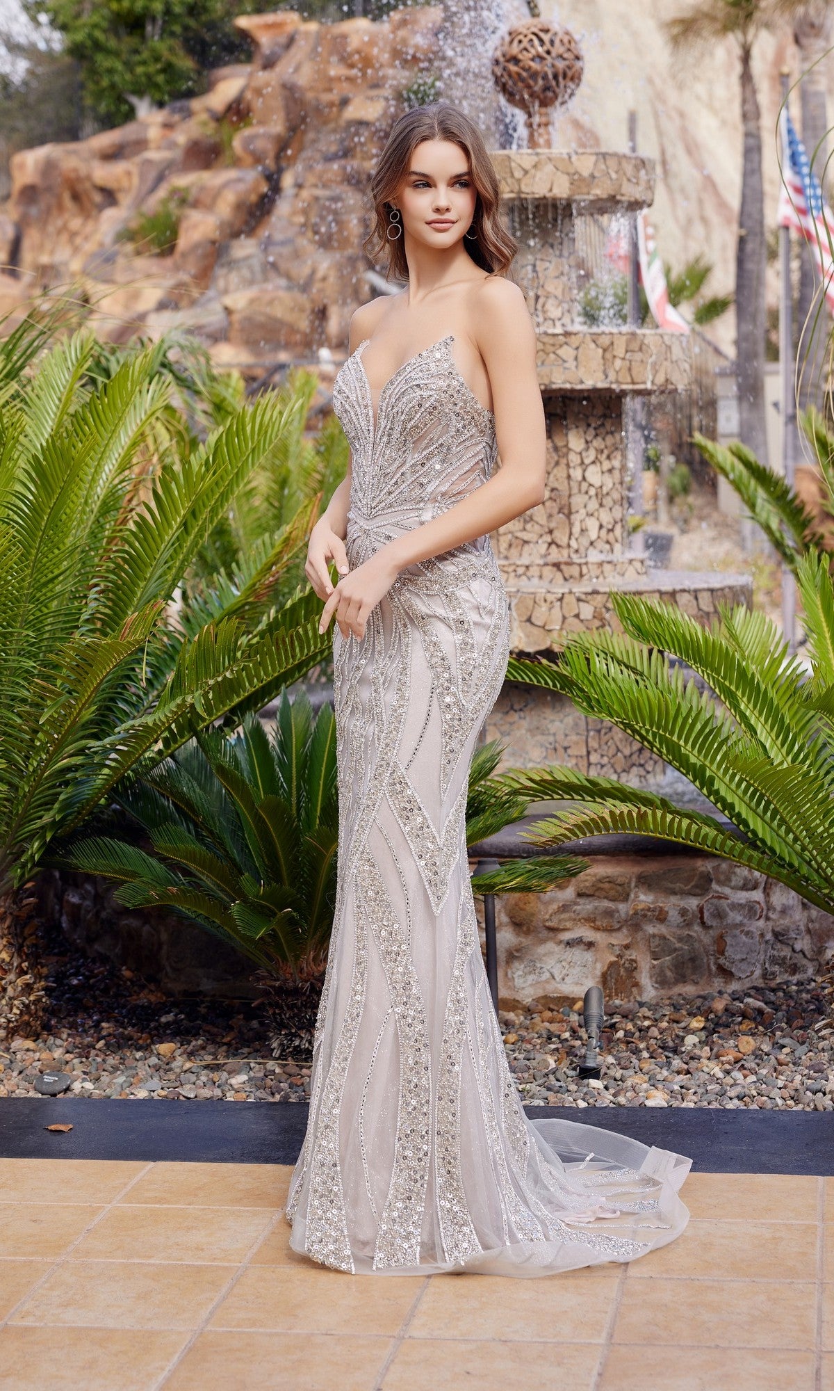 Wearing the Nox Anabel C1833 long prom dress in Silver/Nude, a woman stands outdoors by a stone fountain and lush greenery, her strapless gown shimmering with sequin embellishments.